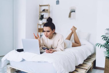 Düşünceli yetişkin kadın, günlük kıyafetlerini giymiş, kredi kartını tutuyor, yatakta yatarken ekrana odaklanıyor ve bulanık arka plandaki açık yatak odasında online alışveriş için dizüstü bilgisayar kullanıyor.