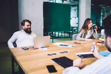 Resmi olarak sakallı yetişkin erkek çalışanlar toplantı masasında dizüstü bilgisayar ve belgelerle otururken modern yaratıcı meslektaşlarıyla sohbet ediyorlar. 
