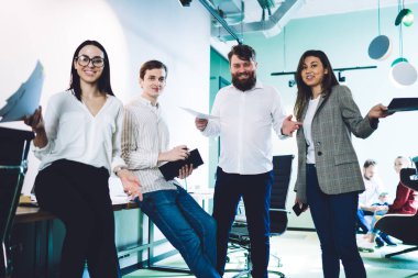 Modern şirkette çalışan, elinde belgelerle çalışan ve gülümseyen kameralara bakan pozitif, çağdaş çalışanlar.