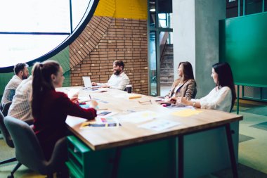 Genç yaratıcı iş adamları büyük toplantı masasında oturmuş gazetelerini dinliyor ve geniş çalışma alanlarında dizüstü bilgisayarları olan erkek liderlerin fikirlerini dinliyorlar.