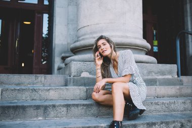 Yoğunlaşmış, uzun saçlı genç bayan cep telefonuyla konuşuyor. Yaz gününde merdivenlerde oturuyor. 