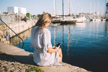 Genç, odaklanmış, uzun saçlı, cep telefonuyla mesajlaşan, yaz günü yatlarıyla göl kenarındaki toprak setinde oturan modern bir kadın. 