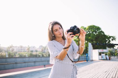 Açık renk elbiseli, siyah noktalı, uzun saçlı genç bir kadın yazın tahta zeminde duran, arka planda büyük bir ağaçla siyah kamerayla fotoğraf çekiyor.