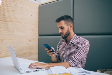 Modern iş yerindeki masada oturan akıllı telefonları kullanarak iş arkadaşlarıyla proje mesajları oluşturmak için dizüstü bilgisayarı kullanan ciddi bir etnik serbest çalışan.