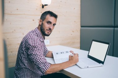 Rahat giyinen, dizüstü bilgisayar kullanan, üniversite projesinde çalışan, kafedeki masada oturup kameraya bakan sakin etnik bir genç.