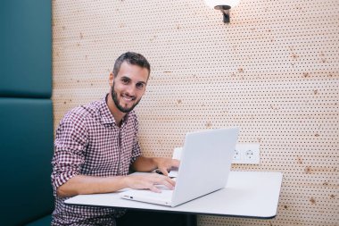 Mutlu gülümseyen, sakallı, sıradan tişörtlü, mavi koltukta oturan ve duvarında lamba olan bir erkek.