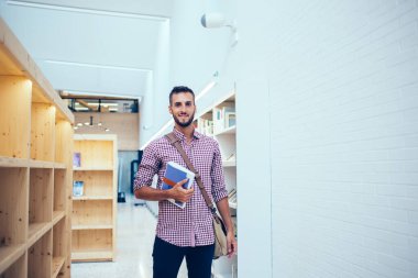 Neşeli, neşeli, zeki, sakallı, çantalı, sıradan tişörtlü, kitap seçen, şehir kitapçısında kitaplarla dolu ahşap rafın önünde duran genç bir erkek.