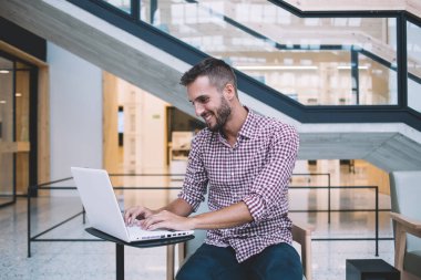 Keyifli etnik gülümseyen adam gündelik kıyafetlerle gezen dizüstü bilgisayarla ofis masasında oturan projenin gelirini kontrol ediyor.