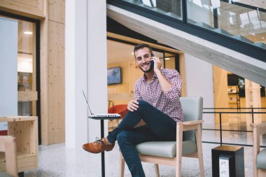 Mutlu gülümseyen, sakallı, normal tişörtlü, cep telefonuyla konuşan yaratıcı iş yerinde dizüstü bilgisayarlı genç adam.