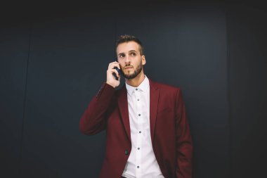 Odaklanmış, beyaz gömlekli, siyah düğmeli, kırmızı ceketli, gri arka planda cep telefonundan arayan ciddi, sakallı bir erkek.