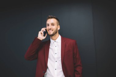 Kırmızı takım elbiseli mutlu sakallı bir erkek, gülümsüyor ve kameraya bakıyor. Siyah duvarın karşısında duruyor ve ofiste akıllı telefondan konuşuyor.