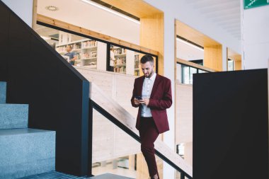 Modern geniş çalışma sahasında merdivenleri çıkarken gülümseyen ve akıllı telefon kullanan modaya uygun kırmızı formalı pozitif genç adam.