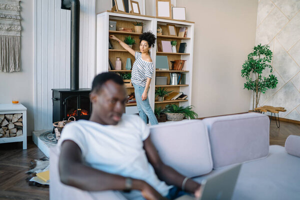 African American man browsing laptop sitting on sofa and looking at camera while ethnic smart ethnic woman taking book from bookshelves in living room