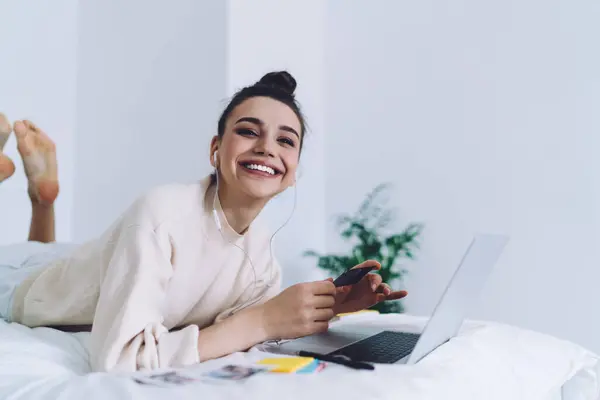 Side view of joyful young woman chilling at home by laptop with earphones holding business card or photo and looking at camera