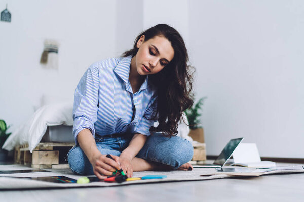 Вдумчивая прилежная женщина с длинными волосами сидит на полу и готовится к подписанию презентаций с зелеными маркерами документов и рисунков 