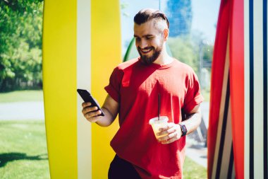 Yakışıklı sakallı genç adam elinde bir fincan taze içecek ve akıllı telefon tutarken güneşli bir günde sörf tahtalarının yanında duruyor ve gülümsüyor.