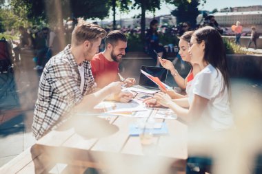 Yazın açık terasta doküman çizelgelerini inceleyen ve masa başında konuşan bir grup zeki genç serbest yazar. 