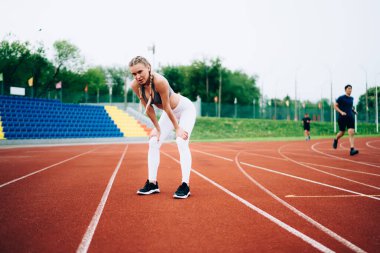 Modern spor giyimli genç, zayıf bir kadın yarış pistinde duruyor. Stadyum koltuklarına karşı antrenmandan sonra dizlerinin üzerine çöküp kameraya bakıyor. 