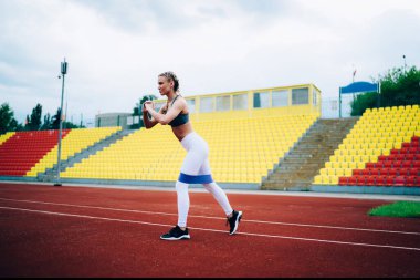 Stadyumda egzersiz yapan kadın sporcu. Ayakları açık stadyumda ayakta duran, elastik direnç bandıyla kalçalarına takılan ve egzersiz yapan genç bayan sporcunun yan görüntüsü.