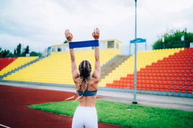 Stadyumda kollarını kaldırıp sıkı antrenman sırasında elastik bantla egzersiz yapan tanınmamış zayıf bir bayanın arka görüntüsü.