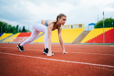 Spor kıyafetleri içinde zayıf, kendine güvenen bayan koşucu antrenman yaparken kırmızı yolda koşmaya başlıyor ve bulanık arka planda stadyuma bakıyor.