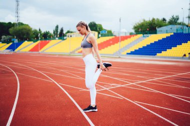 Beyaz taytlı, gri tişörtlü, koşu pistinde duran ve stadyumdaki bacaklarını esneten kadınların yan görüntüsü.