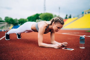 Kendine güveni olan atletik bir kadının kalas egzersizi yapması ve boş tribünlerle stadyumun koşu parkurundaki akıllı telefon ekranına bakması.