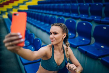 Yukarıdan örülmüş ince bir kadın akıllı telefonuyla selfie çekerken stadyumdaki tribün sandalyesinde oturuyor. 