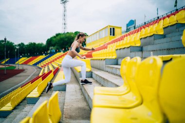 Sportif kıyafetler giyen, bacakları esneyen, stadyumdaki tribünde zıplayan atletik bir kadının yan görünüşü.
