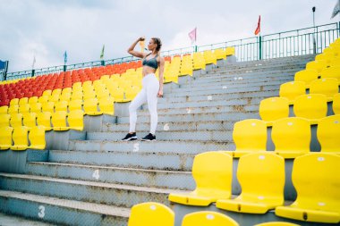 Düşük açılı, modern eşofmanlı, kendine güveni tam bir kadın stadyumda güçlü kollarını gösterirken sayılı basamaklarda dikiliyor.