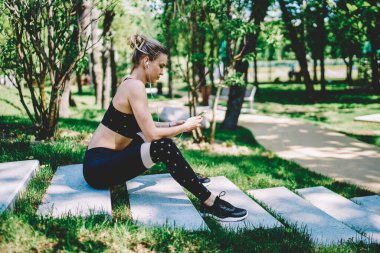 Spor elbiseli, kulaklıklı, telefon dinleyen, parkta merdivenlerde koşmaya hazırlanan aktif bir kadının yan görüntüsü.