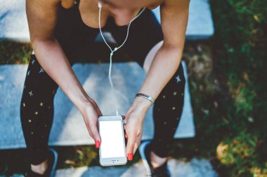 Spor elbiseli, kulaklıklı genç bir bayanın üstünden cep telefonuyla arkadaşlarıyla mesajlaşıp parkta merdivenlerde koşuya hazırlanıyor.