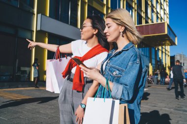 Düşünceli, kese kağıdıyla gezen, açık hava mağazaları arayan, telefon kullanan ve alışveriş yaparken yol gösteren zarif moda kız arkadaşların yan görüntüsü.