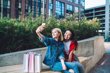 Çok ırklı, şık giyinen genç bayan öğrenciler cep telefonlarıyla selfie çekiyorlar ve caddede bankta otururken gülümsüyorlar. 