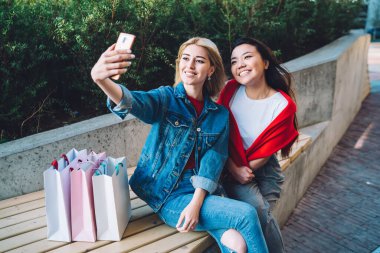 Yukarıdaki neşeli, çekici ve çeşitli kız arkadaşlardan. Parktaki bankta alışveriş yaptıktan sonra birlikte dinlenirken akıllı telefondan selfie çekiyorlar.