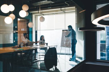 Gündelik kıyafetli meslektaşlar, modern tasarım ofisinde büyük pencereli yeni iş stratejisi sunan patronu dinliyorlar.