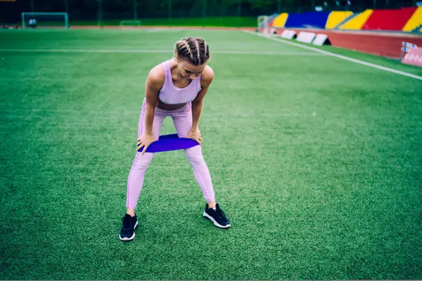 Young sporty fit woman in bright sportswear training with elastic band putting hands on legs standing on green grass of city stadium looking down