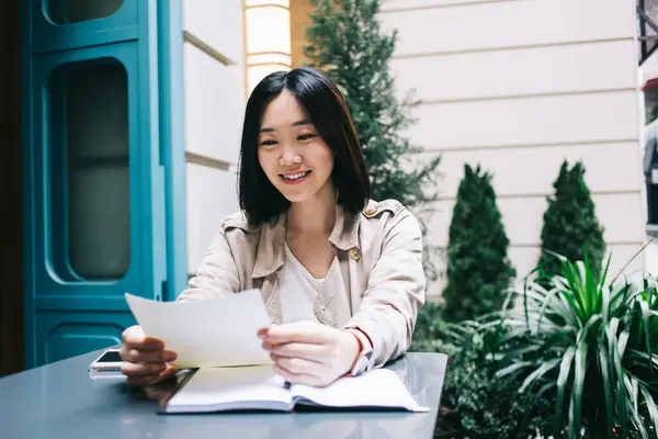 年轻而快乐的亚洲女人 留着一头黑黑的短发 微笑着坐在办公桌前 拿着笔记本 一边在城市街道上看书 — 图库照片