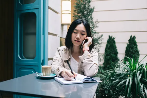 デスクに座ってコーヒーを飲みながらスマートフォンで話す短い黒髪の集中した若い女性 — ストック写真