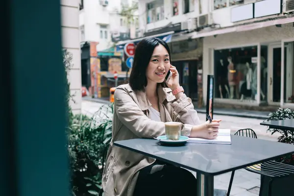 侧视图年轻的黑发女子坐在办公桌前喝咖啡 一边用智能手机聊天 一边看着相机 一边在路上的笔记本上写字 — 图库照片