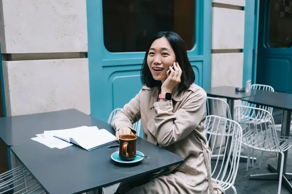 Gelukkig Aziatische Vrouw Trench Spreken Smartphone Terwijl Zitten Aan Tafel — Stockfoto