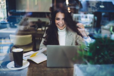Öğrenmek ve kameraya gülümsemek için bilgi kitabı ve dizüstü bilgisayarla masaüstünde oturan mutlu hipster öğrencisinin portresi, uzaklık planıyla uzaktan çalışan başarılı kadın serbest yazar