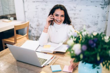 İçerikten mutlu, uzun siyah saçlı, beyaz kazakla gülümseyen, cep telefonuyla sohbet eden yaratıcı iş yerinde dizüstü bilgisayarlı ve defterli bir kadın. 