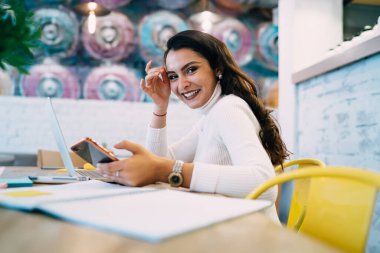Kafeteryada dizüstü bilgisayar ve not defteri ile ders hazırlarken ve kameraya bakarken günlük giysiler içinde gülümseyen esmer kadının yan görüntüsü.