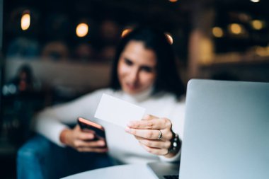 Gündelik giysiler içindeki genç bayan akıllı telefon kullanıyor. Kartvizitini gösteriyor. Kafede arka planda bulanık bir şekilde oturuyor.