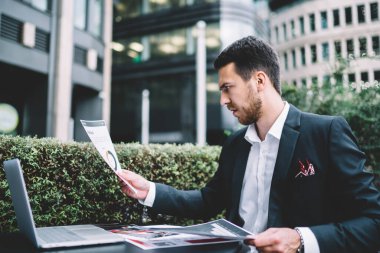 Kafe masasında bilgisayarla uzaktan çalışma sırasında resmi takım elbise içinde kurumsal kağıt diyagramları üzerinde çalışan konsantre olmuş zeki bir yönetici.