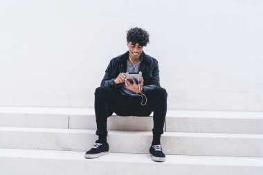 Afro-Amerikalı genç yakışıklı adam merdivenlerde gülümsüyor, tablet üzerinde mesaj yazıyor ve kulaklıkla beyaz duvara karşı müzik dinliyor.