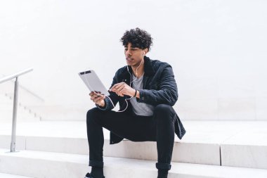 Siyah elbiseli genç Afro-Amerikan erkek merdivenlerde oturup tabletten müzik seçiyor beyaz arka planda ekrana bakıyor.