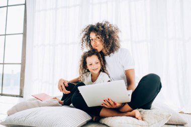 Gündelik giysiler içinde etnik anneyi önemseyen, dizüstü bilgisayar seyreden, ışık stüdyosunda yastıkların üzerinde gülümseyen, kıvırcık saçlı, sevimli bir kızla oturan.