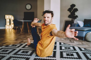 Kırmızı tişörtlü, mavi pantolonlu, el ve bacağını halının üzerinde uzatan genç bir erkek. 